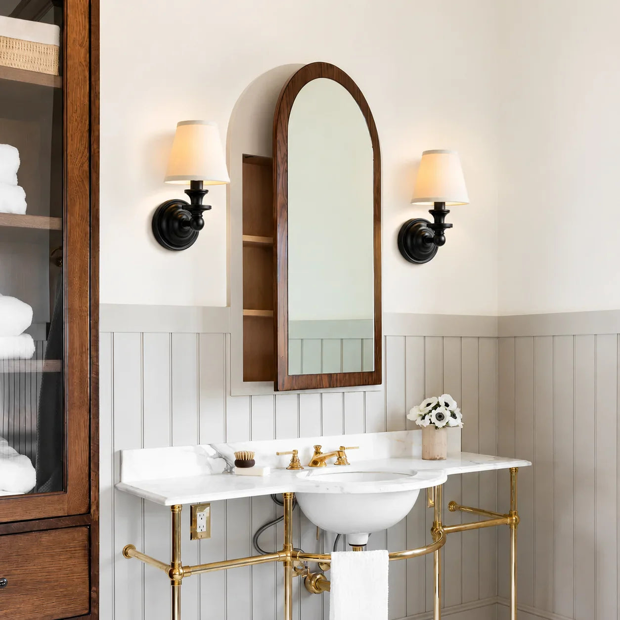 A bathroom vanity scene with a white pedestal sink, brass pipe legs, a white marble countertop, a round wood-framed mirror, two black wall sconces with cream shades, and gray wainscoting on the wall.