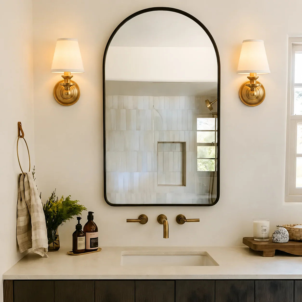 An arched rectangular wall mirror with a thin black frame hangs above a bathroom vanity. The scene includes two brass sconces, a brass faucet and handles, a cream countertop, a towel on a ring, and a window on the right.