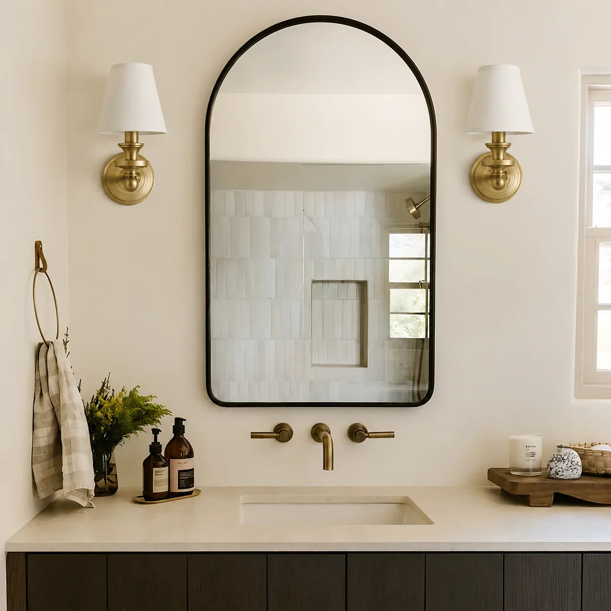 An arched wall mirror with a thin black frame hangs above a bathroom vanity with a cream countertop and matte brass fixtures. Two matching brass sconces with white shades flank the mirror, and toiletries sit on the counter.