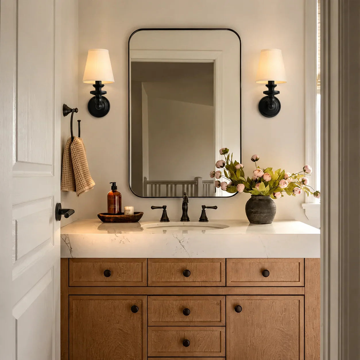 A styled bathroom vanity featuring a light oak wood cabinet, a white marble countertop, a black faucet, a large rectangular mirror with a thin black frame, and two black wall sconces with white shades on either side.