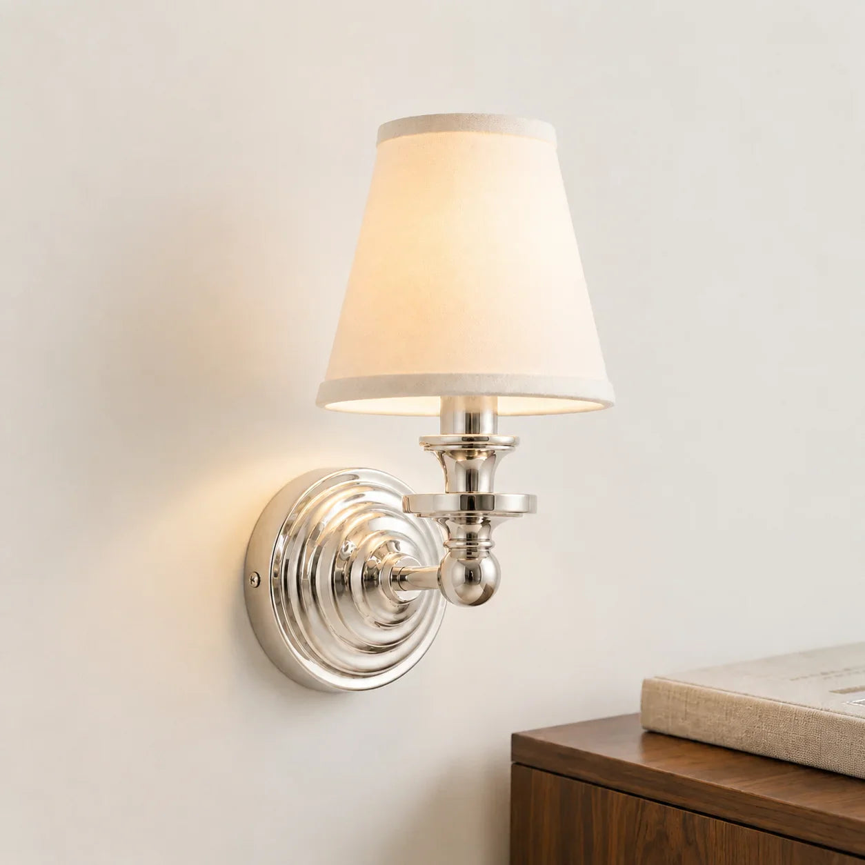A wall-mounted sconce lamp with a warm white fabric shade and a polished silver metal base, shown lit against a light neutral wall above a wooden surface.