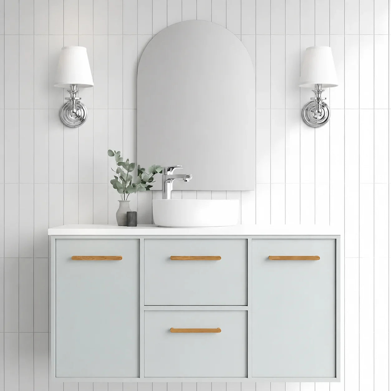 A modern bathroom vanity set featuring a white vessel sink, a light gray floating cabinet with natural wood handles, a large arched mirror, and two wall-mounted sconces against a white tiled wall.