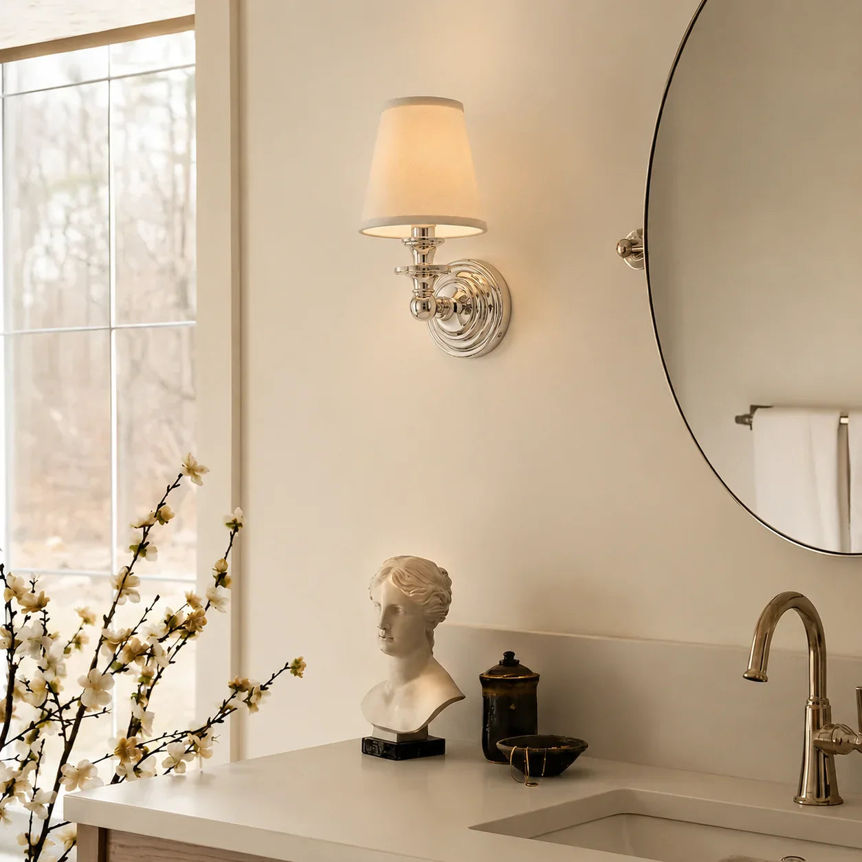 A wall-mounted sconce with a warm white fabric shade and polished silver metal base, installed above a bathroom vanity with a mirror, faucet, and decorative bust.