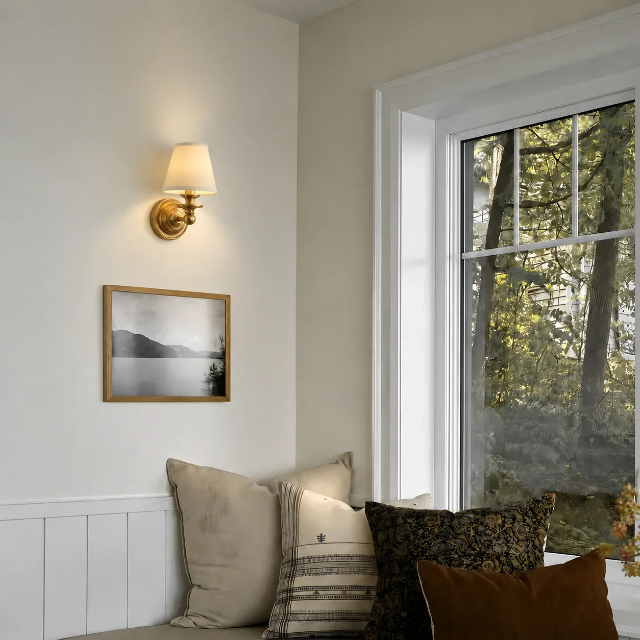 A brass wall sconce with a warm white fabric shade is mounted on a beige wall above a framed black-and-white landscape print and a cushioned bench with neutral and brown pillows, next to a window showing trees outside.