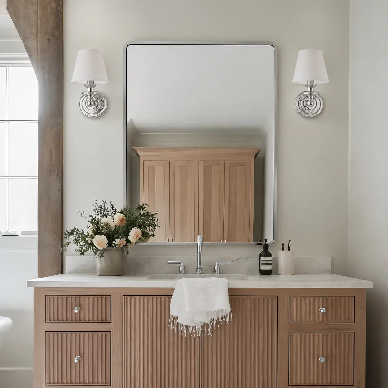 A modern bathroom vanity featuring a light wood cabinet with ribbed doors, a white marble countertop, a large rectangular mirror, and two matching wall sconces on a beige wall.