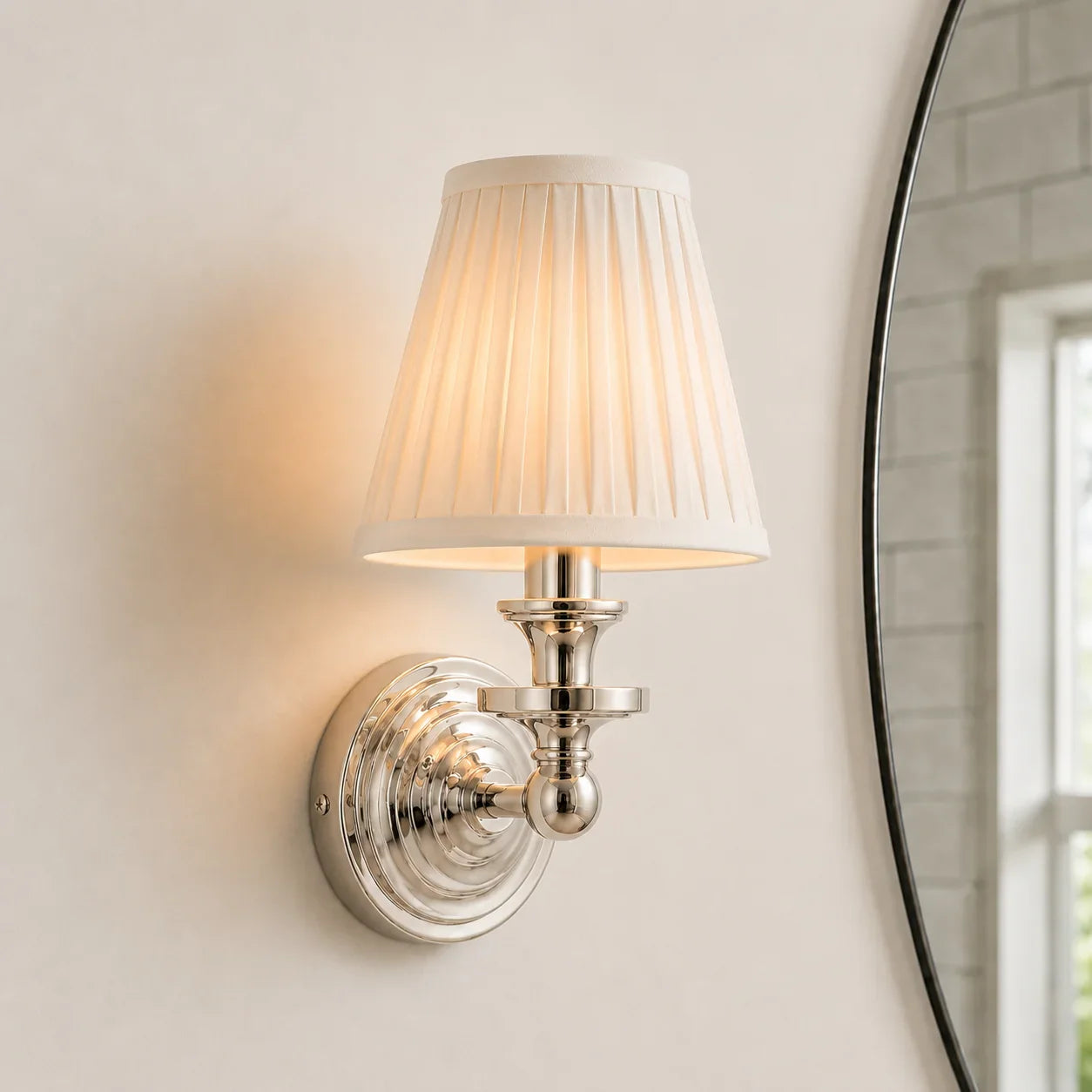 A wall-mounted sconce with a pleated white fabric shade and a polished silver metal base, shown lit against a warm off-white wall.