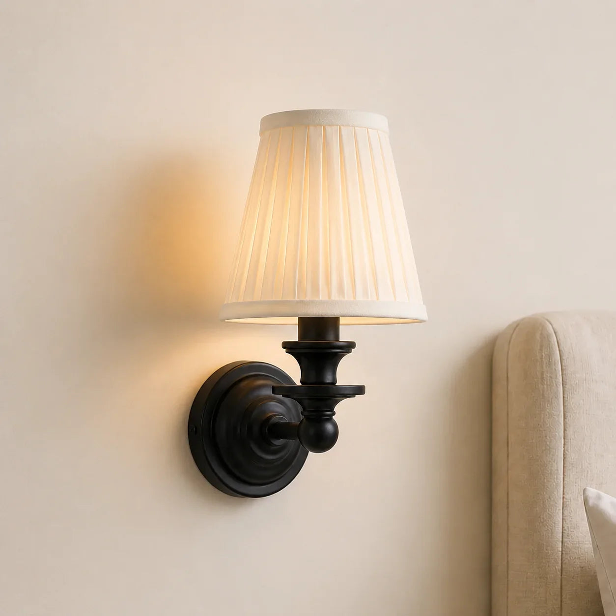 A wall-mounted sconce with a black metal bracket and a pleated off-white fabric shade, shown lit against a beige wall with part of a upholstered chair visible on the right.