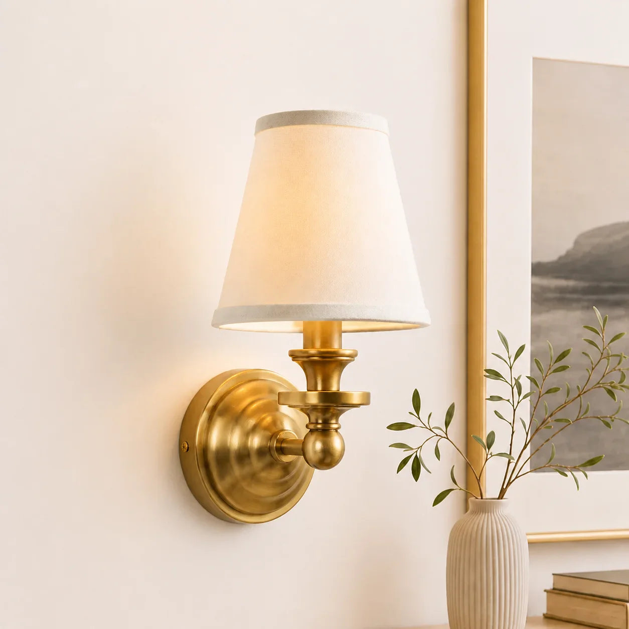 A brass wall sconce with a tapered off-white fabric shade, mounted on a beige wall next to a framed print and a small vase with greenery.