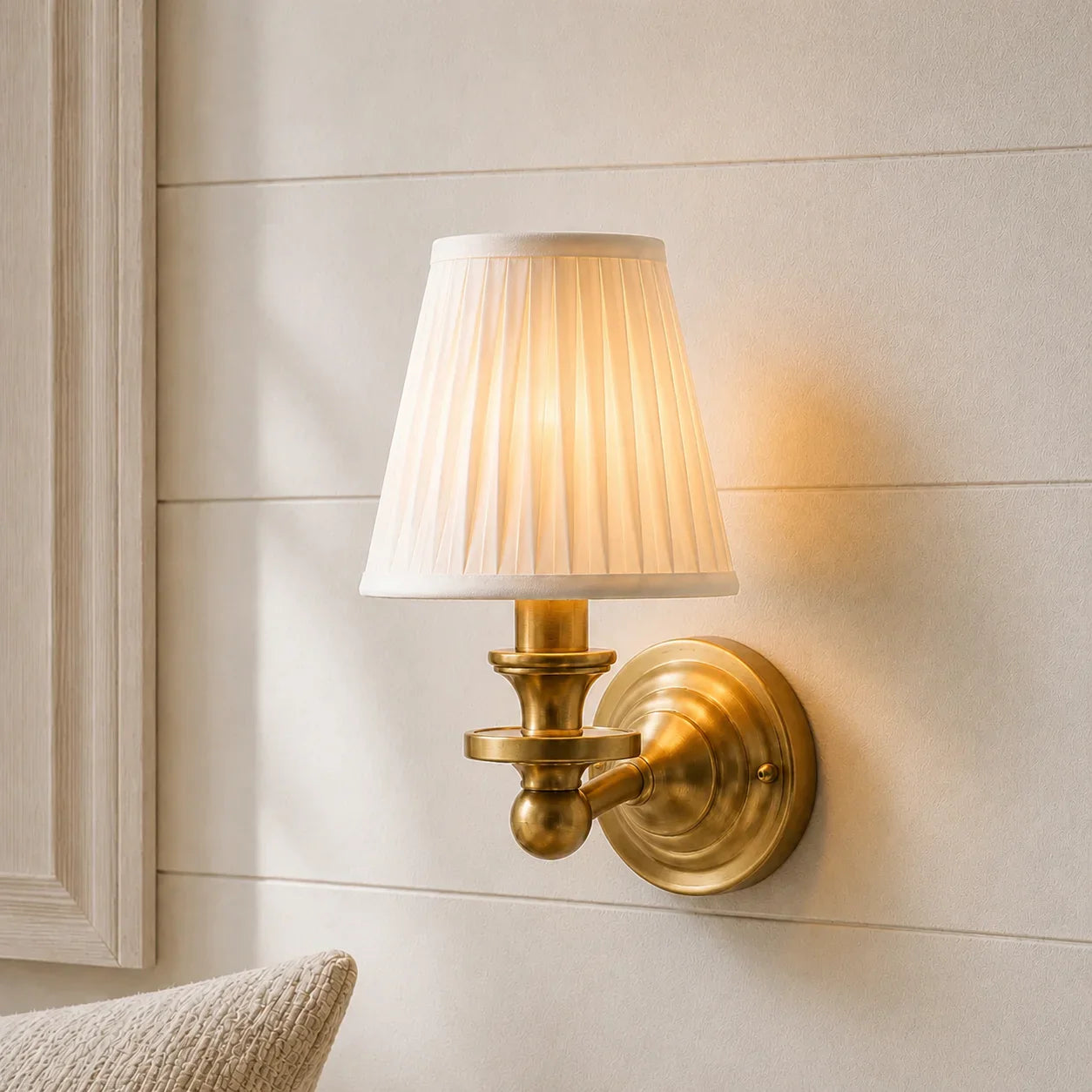 A wall-mounted sconce with a pleated white fabric shade and an antique brass/gold metal fixture, shown lit against a beige shiplap wall.