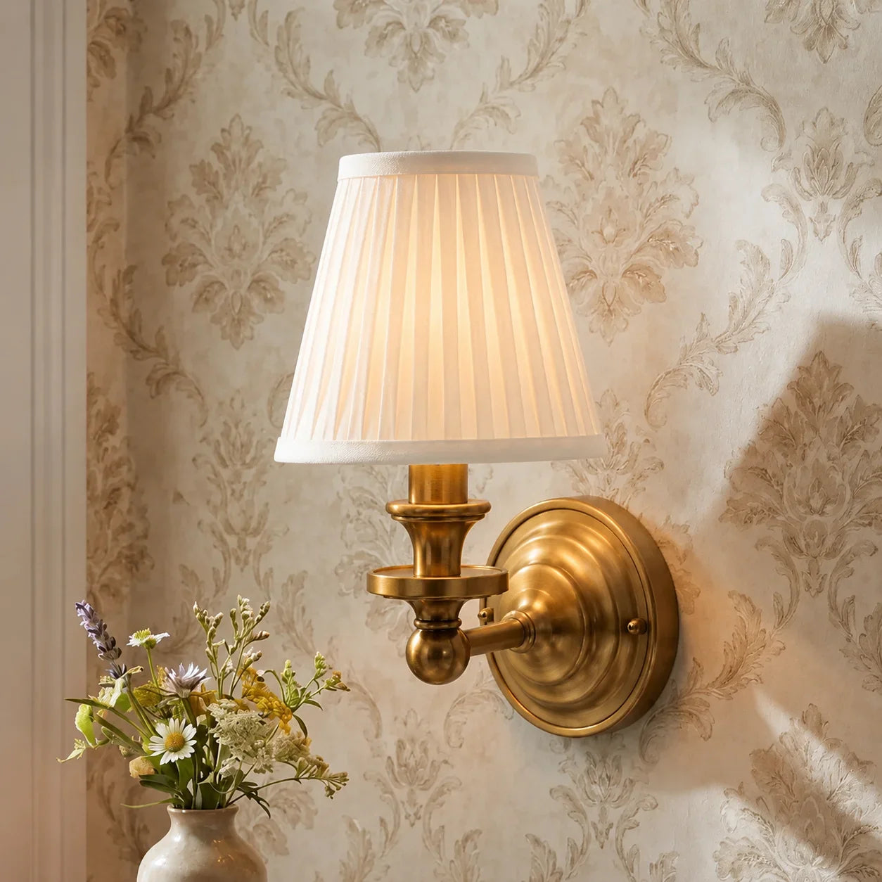 A classic brass wall sconce with a pleated white fabric shade, mounted on a beige damask-patterned wallpapered wall next to a vase of flowers.