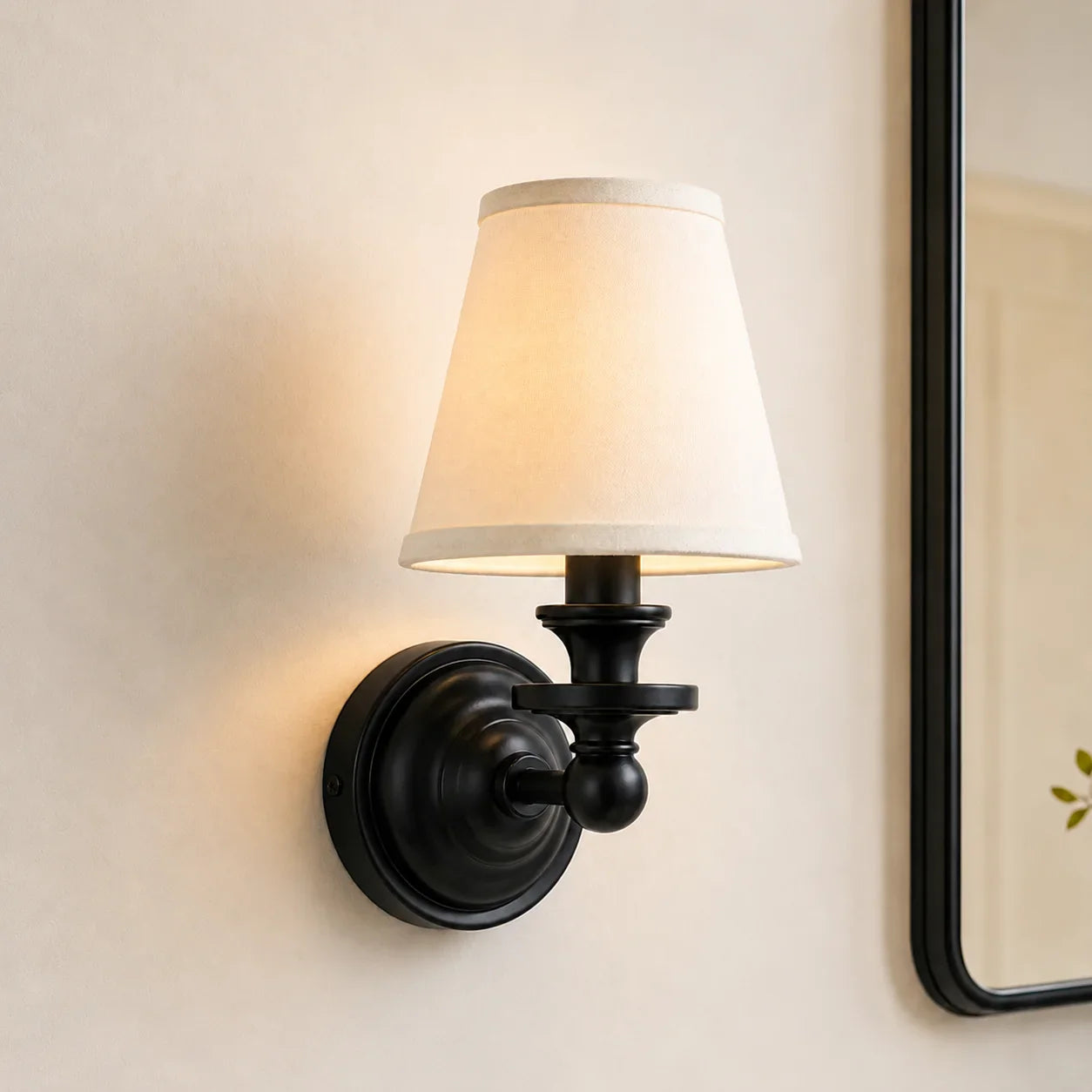 A single wall sconce light fixture with a black metal mount and a tapered off-white fabric shade, shown lit against a warm beige wall.