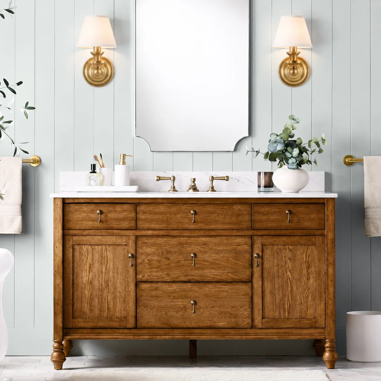 A double-sink bathroom vanity in warm medium brown wood with brass hardware, set against a light blue paneled wall. The vanity has a white marble countertop with a white double basin, matching brass faucets, and a large rectangular mirror above.