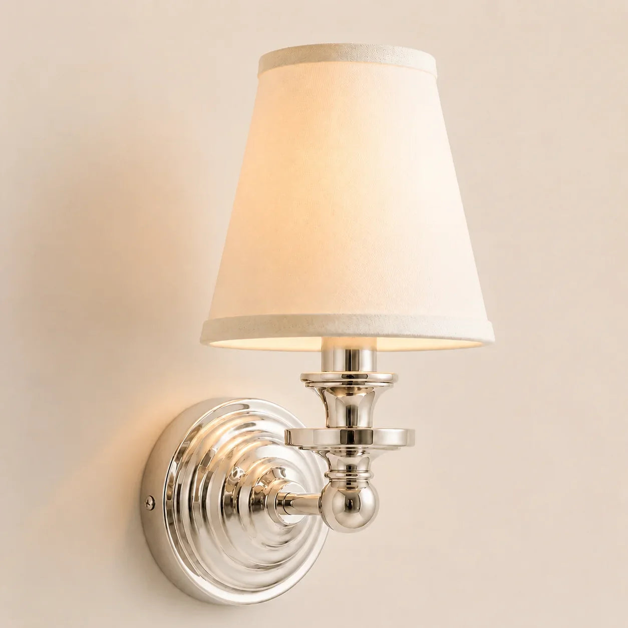 A wall-mounted sconce with a warm white fabric shade and a polished silver metal base, shown lit against a plain beige wall.