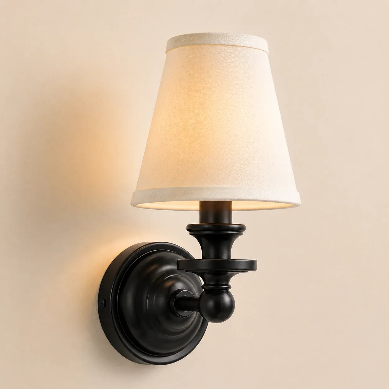 A wall-mounted sconce light fixture with a black metal wall plate and arm, featuring a warm white fabric drum shade, shown lit against a beige wall.