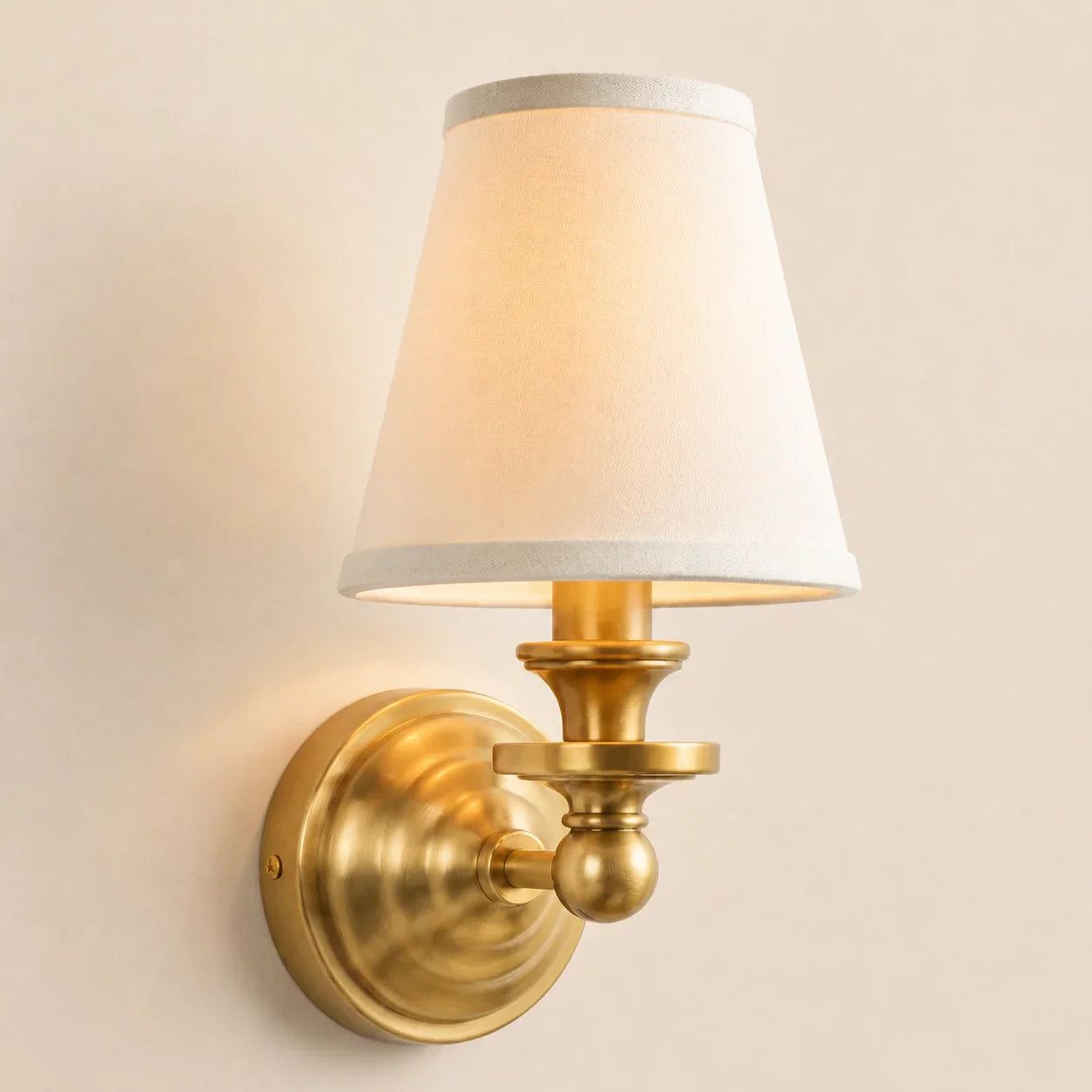 A wall-mounted sconce with a warm brass/gold metal fixture and a tapered off-white fabric shade, shown lit against a plain beige wall.