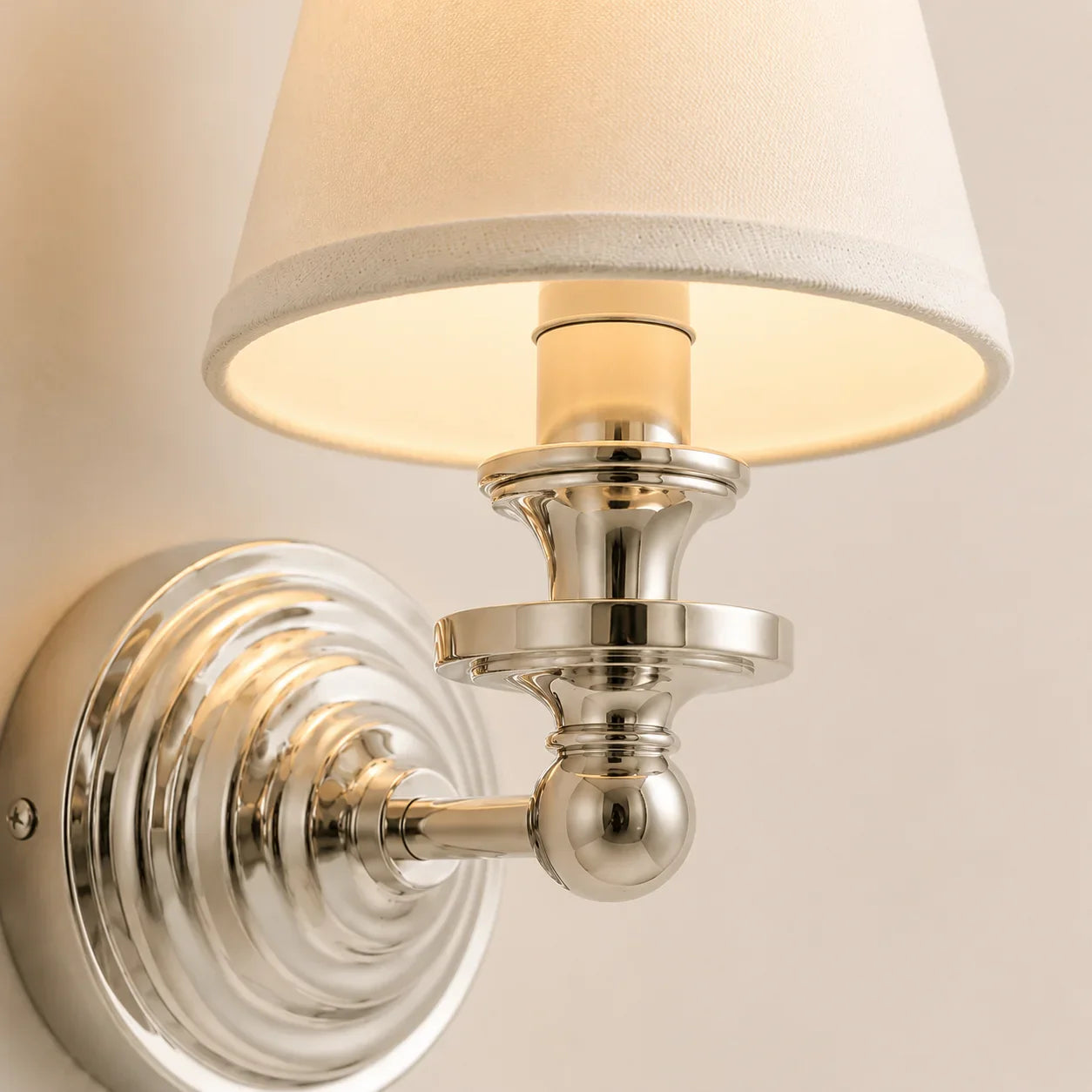 A close-up of a wall-mounted sconce with a warm white fabric drum shade and polished silver metal hardware against a light beige wall.