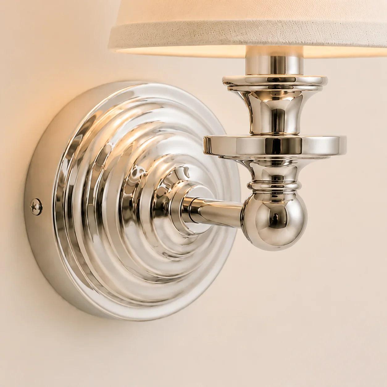 A close-up of a wall-mounted sconce with a polished silver metal finish and a warm beige fabric shade, shown lit against a light beige wall.