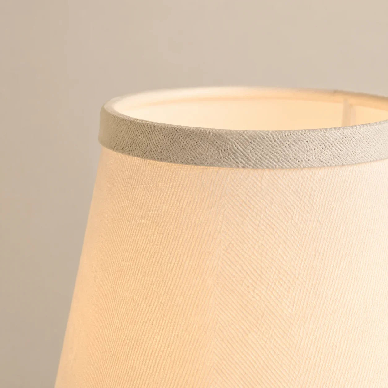 Close-up of a fabric lampshade in a warm cream or beige tone with a textured woven band around the top edge, shown against a plain light gray background.