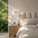 Neatly made bed with beige bedding and nightstand in a room with a large window showing greenery outside.