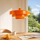 A modern three-tiered orange pendant light hanging over a wooden dining table with oranges, a white mug, and an open magazine, set against a warm beige wall and a blurred window view of green trees and blue sky.