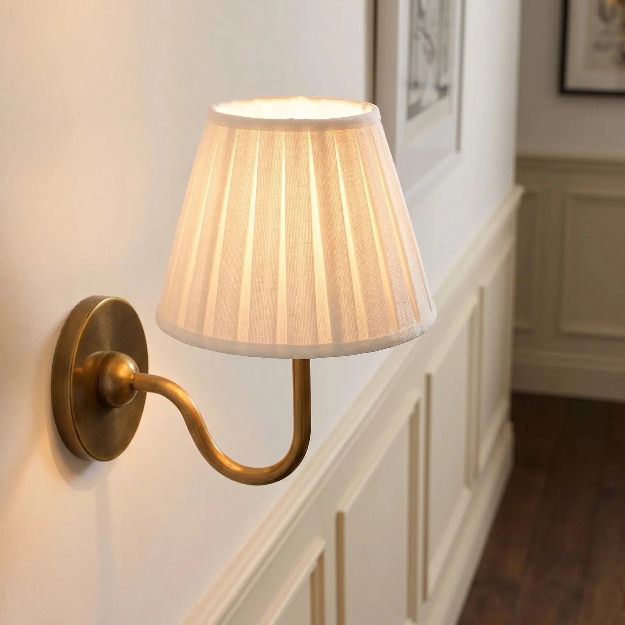 Wall-mounted lamp with pleated white shade and brass arm on a wall.