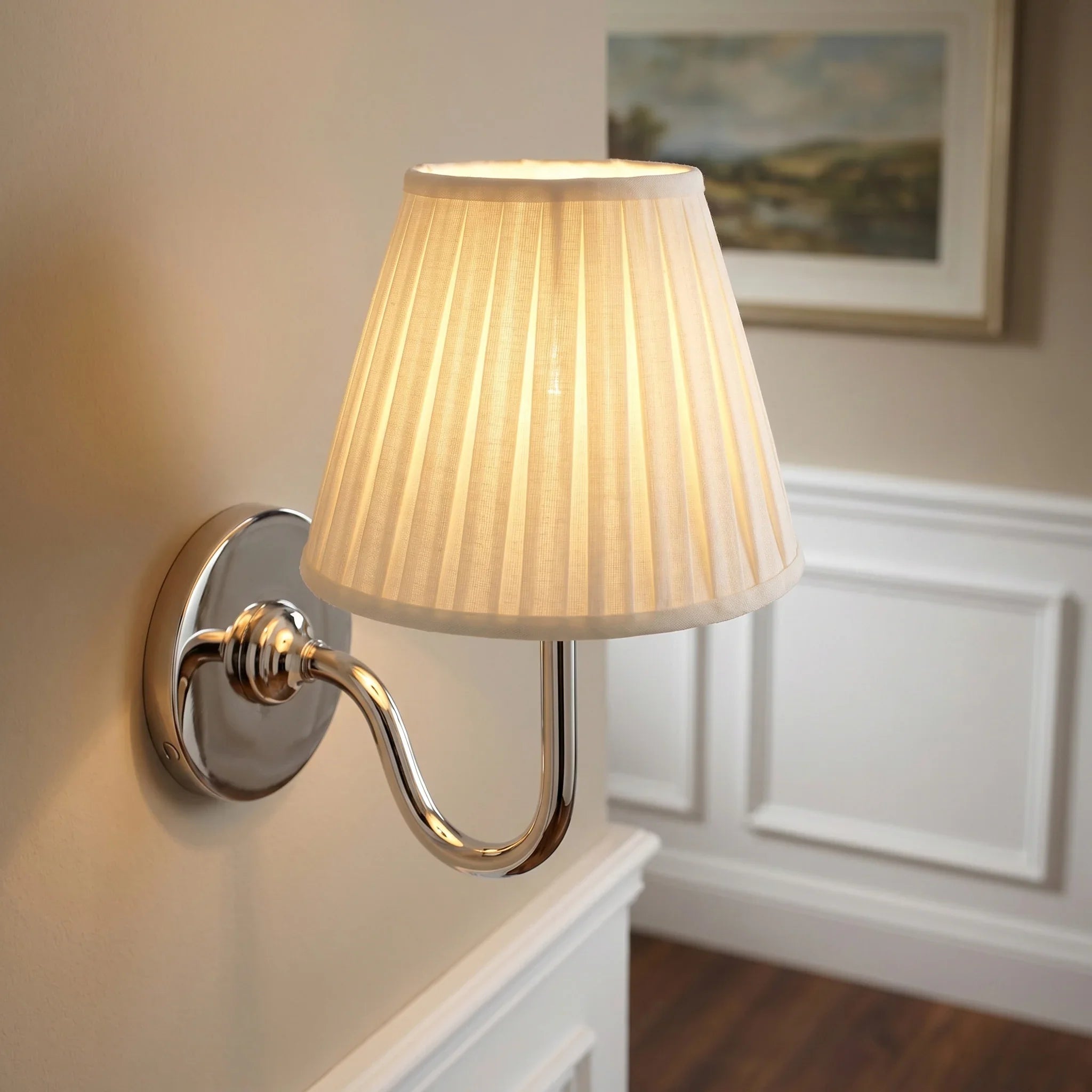 Wall-mounted lamp with pleated shade on a wall with decorative molding and a painting.
