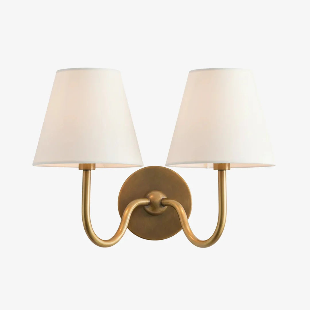A double-light wall sconce with a warm brass-finished metal frame and two tapered off-white fabric shades, shown against a plain white background.