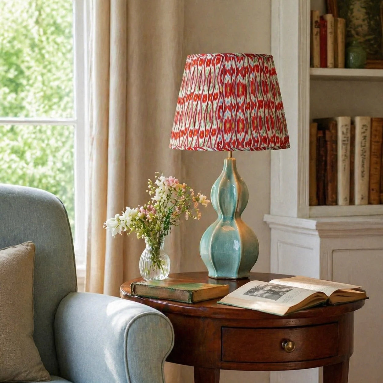 A table lamp with a turquoise-green sculptural base and a red, white, and orange patterned drum shade sits on a dark wood side table in a cozy room setting next to a light blue armchair.