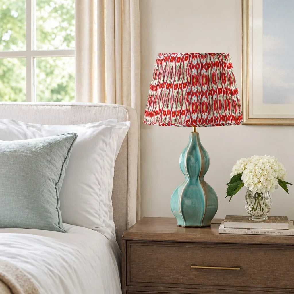 A table lamp with a turquoise ceramic base and a red, white, and gray patterned drum shade, styled on a wooden nightstand next to a bed in a bedroom.