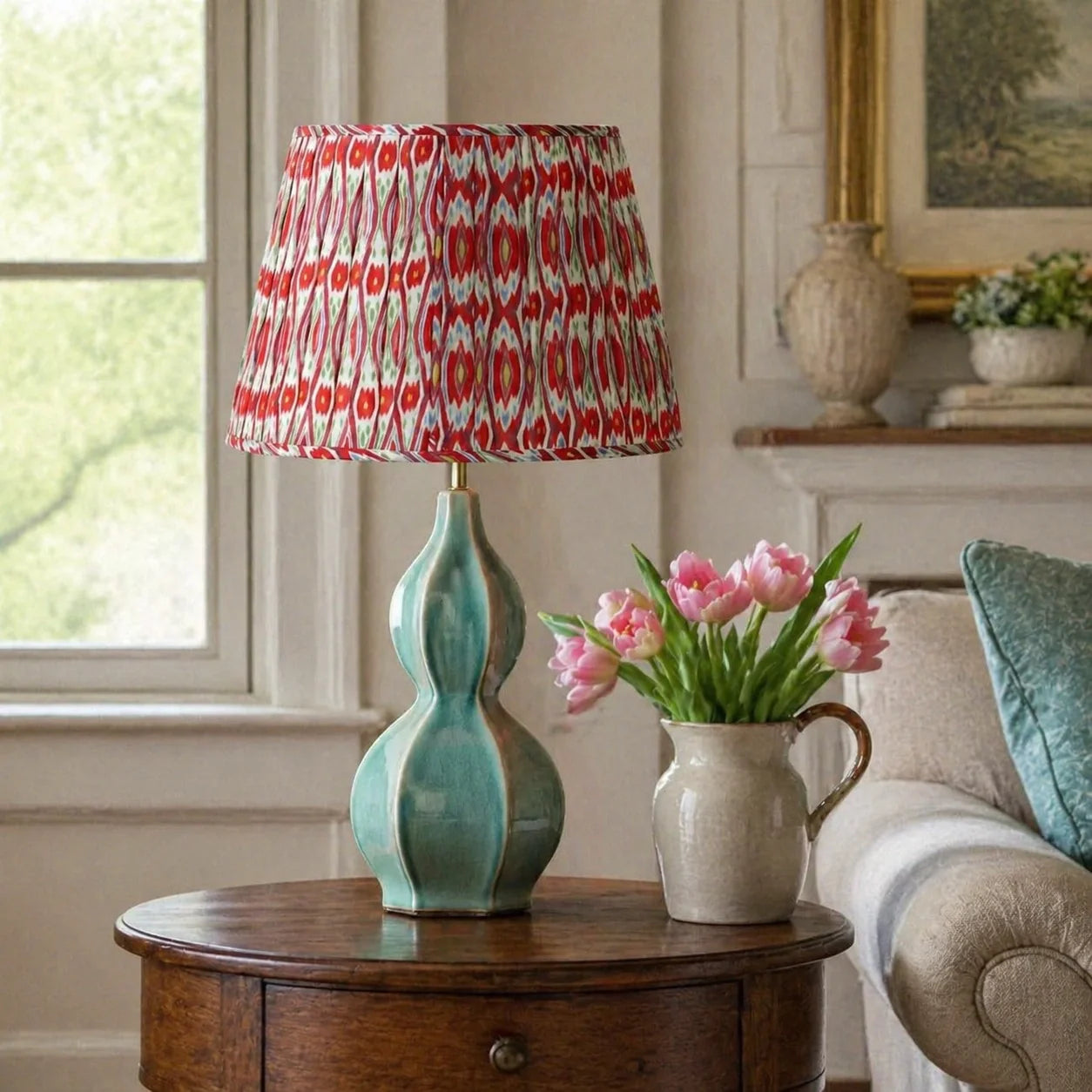 A table lamp with a turquoise-green ceramic gourd-shaped base and a red, white, and black patterned pleated drum shade, styled on a wooden side table next to pink tulips in a cozy living room.