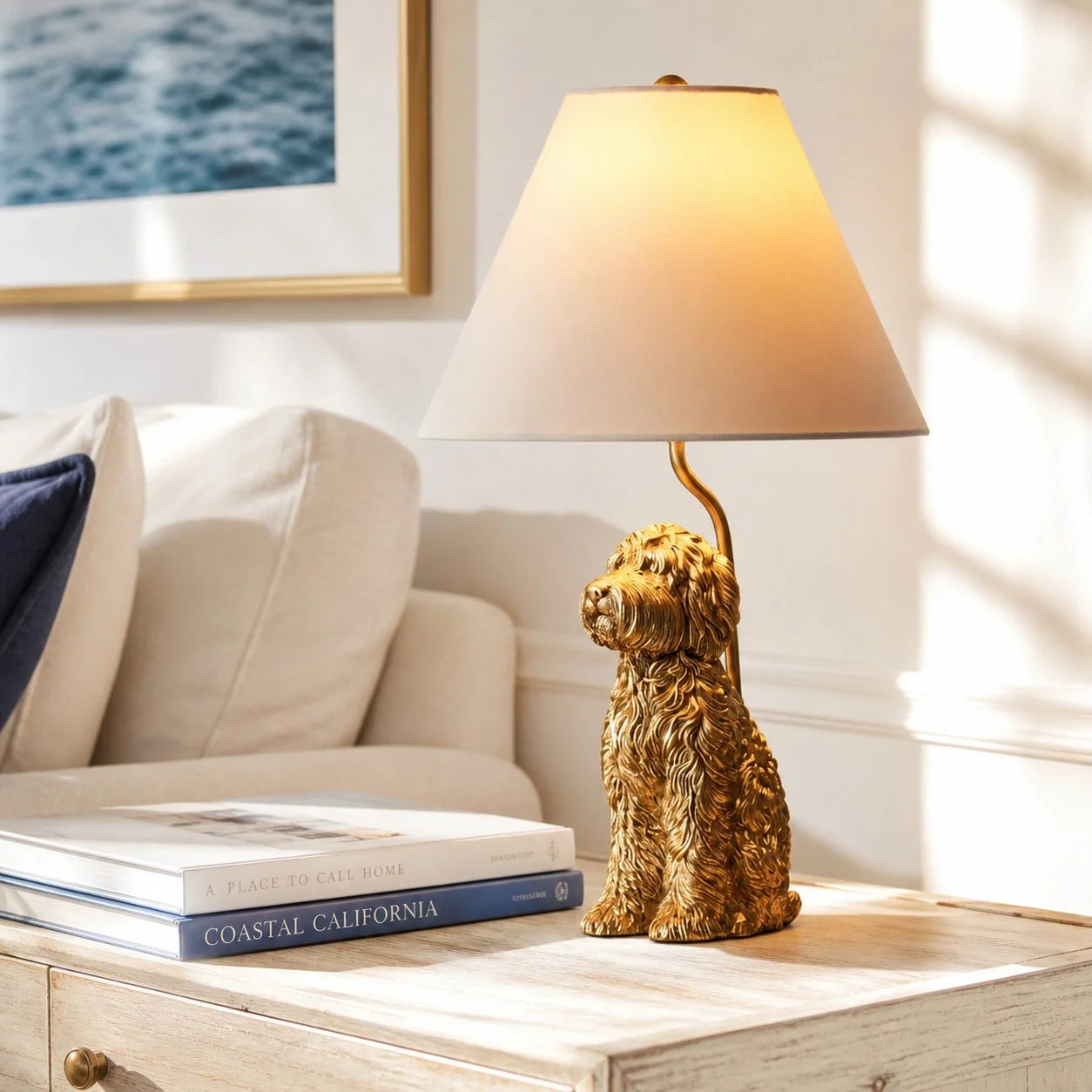 A decorative table lamp shaped like a golden dog sitting on a light wood side table in a bright living room. The lamp has a warm beige conical shade and glows with a warm yellow light.