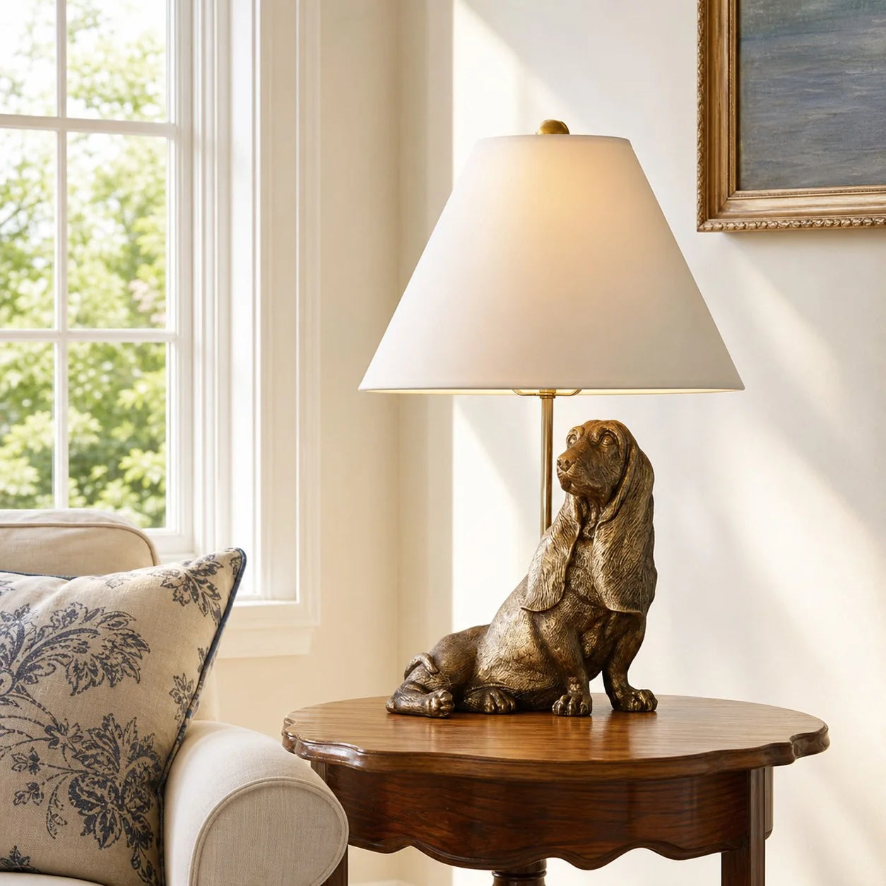 A table lamp featuring a bronze-colored dog figurine base and a white conical shade, placed on a wooden side table in a bright living room next to a window and patterned pillow.