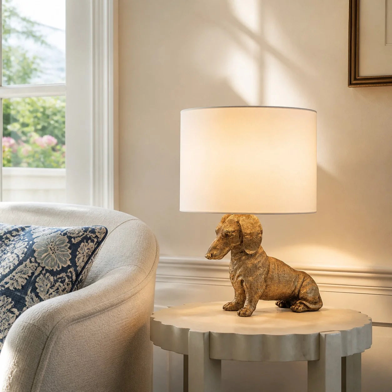 A table lamp with a warm-toned dachshund figurine base and a white drum shade, styled on a round side table next to a beige armchair in a softly lit room.