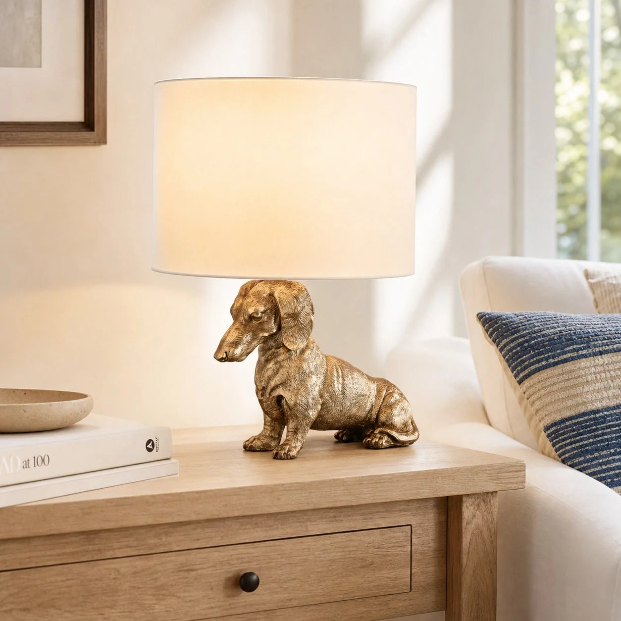 A table lamp with a metallic gold dachshund figurine base and a warm cream-colored drum shade, styled on a wooden side table in a cozy living room setting.