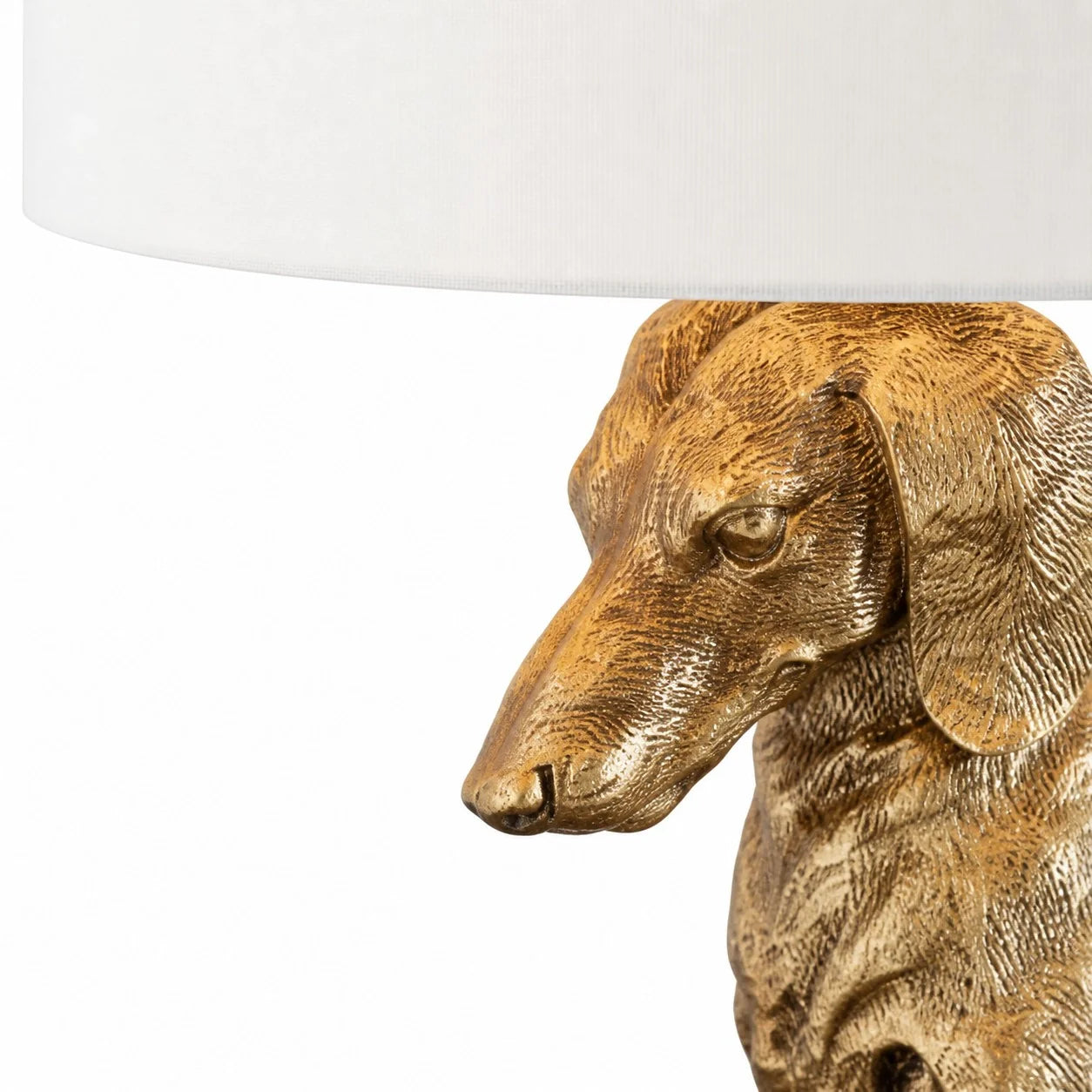 A close-up of a sculptural table lamp featuring a metallic gold dachshund dog head and neck under a white drum shade, set against a plain white background.