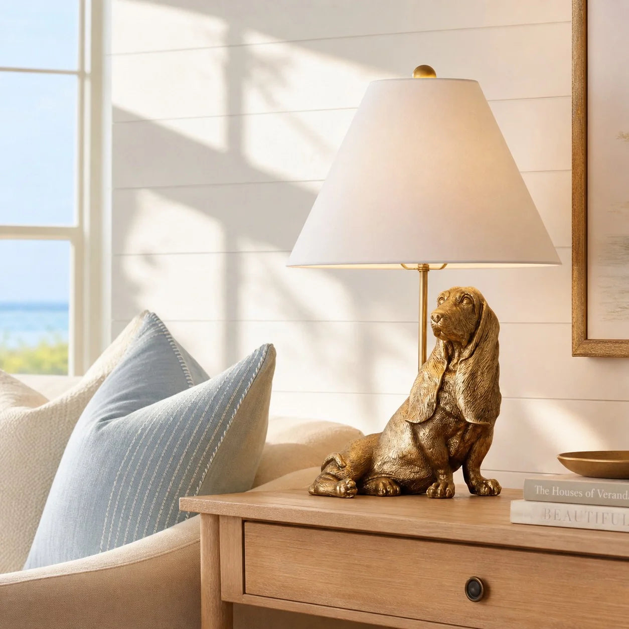 A decorative table lamp shaped like a sitting golden retriever dog in metallic gold, placed on a light wood bedside table next to a cream armchair with blue pillows in a sunlit room.