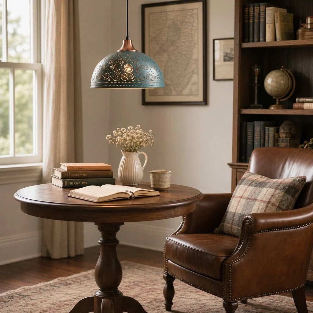A rustic blue-green pendant lamp with gold detailing hangs over a round wooden table in a cozy, sunlit study room with a leather armchair, books, and framed art.